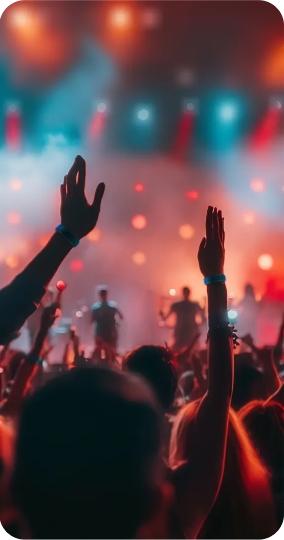 A crowd of people with raised hands at a concert, illuminated by colorful stage lights in a vibrant atmosphere