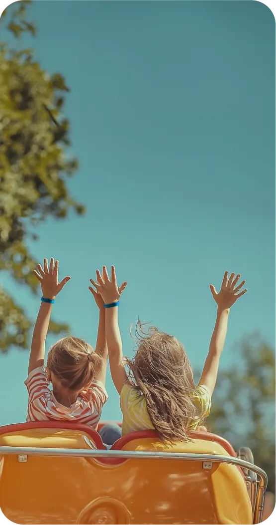 Two girls with their hands raised riding a roller coaster on a sunny day, with a view of the sky and trees