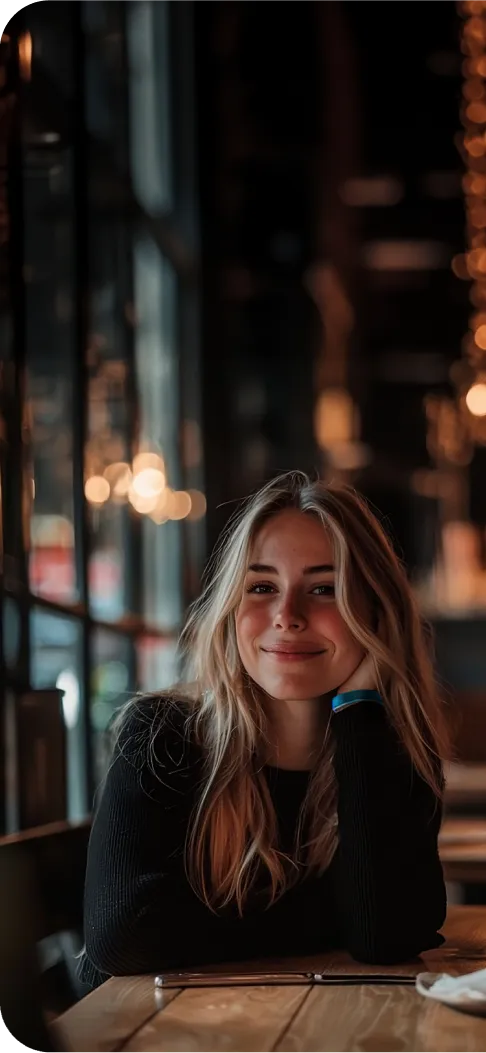 A smiling woman in a black sweater sitting at a wooden table in a cozy café, illuminated by warm lighting
